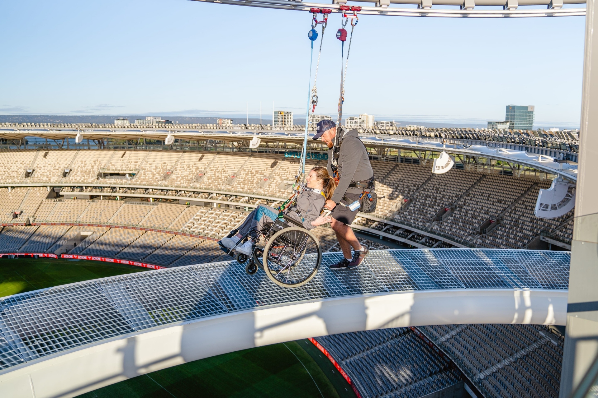 Eine Frau in einem Rollstuhl, die einen Gurt trägt und bei The Ozone über dem Optus Stadium in Perth an einem dramatischen Abgrund entlanggeführt wird, Westaustralien © Tourism Australia