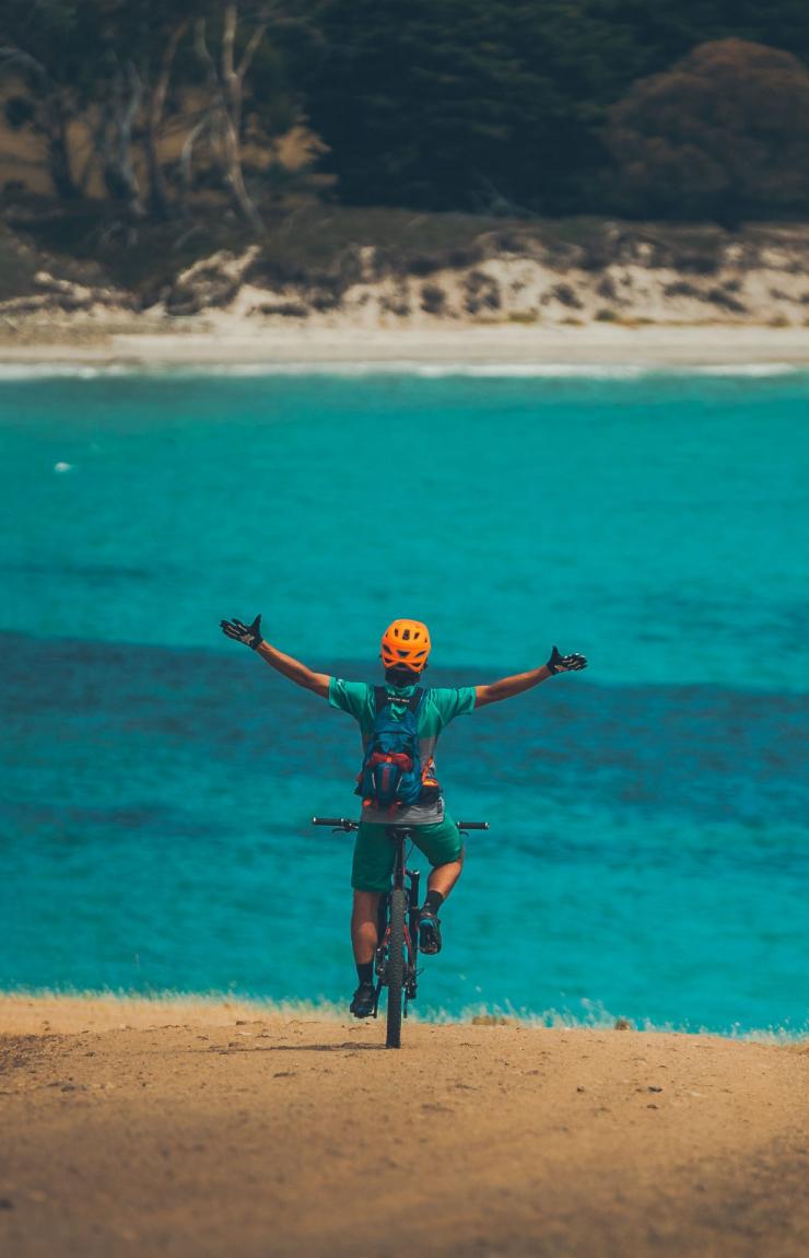 Person auf einem Mountainbike, die die Arme in die Luft hebt und über den strahlend blauen Ozean im Maria Island National Park, Tasmanien, blickt © Matt Staggs