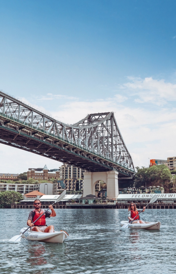 Ein Mann und eine Frau fahren mit Riverlife Kajak auf dem Brisbane River unter der Story Bridge mit den Howard Smith Wharves im Hintergrund in Brisbane, Queensland © Tourism and Events Queensland