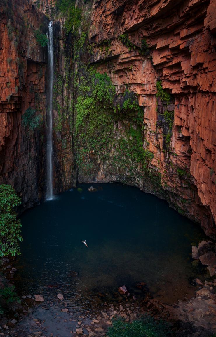 Luftaufnahme einer Person, die sich auf dem Rücken in einem tiefblauen Naturpool treiben lässt, umgeben von hoch aufragenden roten Felswänden, die mit Moos bedeckt sind und von denen ein schmaler Wasserfall stürzt, Emma Gorge, El Questro Wilderness Park, Kimberley, Westaustralien © Tourism Australia