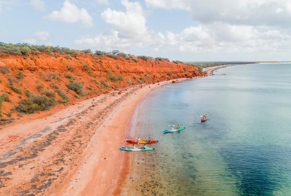 Luftaufnahme einer Gruppe von Menschen, die während einer Tour mit Wula Gura Nyinda Eco Cultural Adventures neben leuchtend orangefarbenen Klippen in klarem blauem Wasser Kajak fahren, Coral Coast, Westaustralien © Tourism Western Australia
