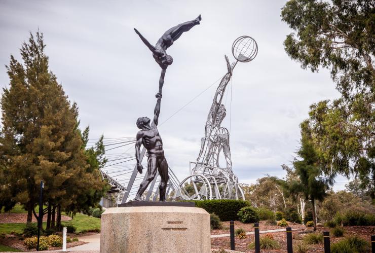 A statue in the foreground with two gymnasts holding hands and a larger statue in the background of a person in a wheelchair reaching for a basketball on a property surrounded by trees at Australian Institute of Sport, Canberra, Australian Capital Territory © Richard Poulton/VisitCanberra A statue in the foreground with two gymnasts holding hands and a larger statue in the background of a person in a wheelchair reaching for a basketball on a property surrounded by trees at Australian Institute of Sport, Canberra, Australian Capital Territory © Richard Poulton/VisitCanberra