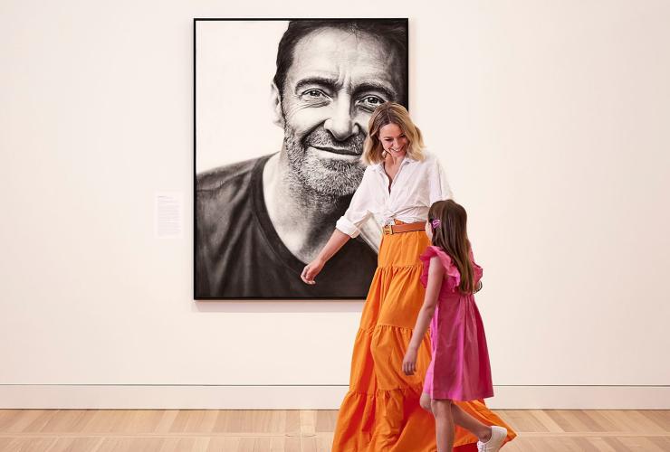 A mother and daughter wearing bright pink and orange clothes walking past a black and white painting of a man smiling against a blank wall at the National Portrait Gallery, Canberra, Australian Capital Territory © Crux for VisitCanberra A mother and daughter wearing bright pink and orange clothes walking past a black and white painting of a man smiling against a blank wall at the National Portrait Gallery, Canberra, Australian Capital Territory © Crux for VisitCanberra