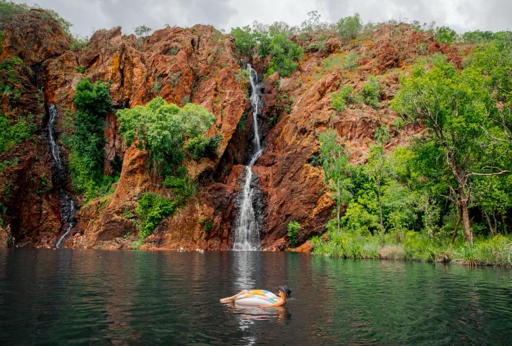 A person laying on a floating device in a waterhole with tall, thin waterfalls cascading in from a red rock cliff sprinkled with green trees at Wangi Falls, Litchfield National Park, Northern Territory © Tourism NT