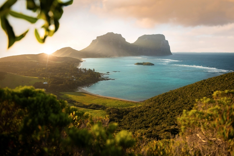 A tree-covered island with bright blue ocean and two towering mountains in the distance at Mount Lidgbird and Mount Gower, Lord Howe Island, New South Wales © Tom Archer