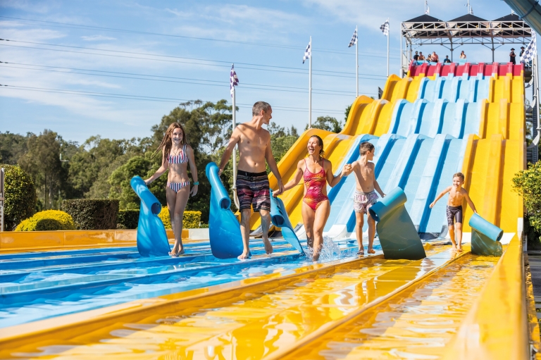 A family smiling and walking along the bottom of a yellow, blue and red slide at Wet ‘N’ Wild waterpark in the Gold Coast, Queensland © Destination Gold Coast A family smiling and walking along the bottom of a yellow, blue and red slide at Wet ‘N’ Wild waterpark in the Gold Coast, Queensland © Destination Gold Coast