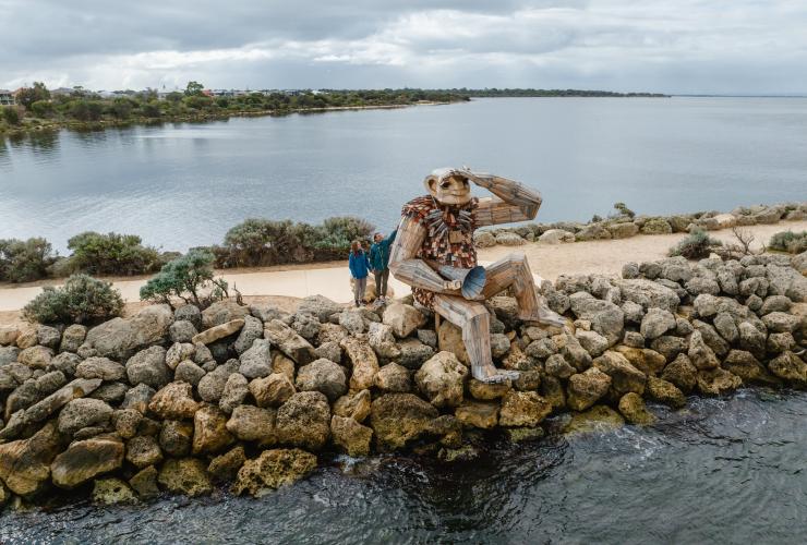 Aerial view over a rock-lined pier over a tranquil waterway, with two people standing beside a giant sculpture that appears to be sitting on the edge and looking out to sea at Seba's Song Giant, Giants of Mandurah by Thomas Dambo, Mandurah, Western Australia © Visit Mandurah Aerial view over a rock-lined pier over a tranquil waterway, with two people standing beside a giant sculpture that appears to be sitting on the edge and looking out to sea at Seba's Song Giant, Giants of Mandurah by Thomas Dambo, Mandurah, Western Australia © Visit Mandurah