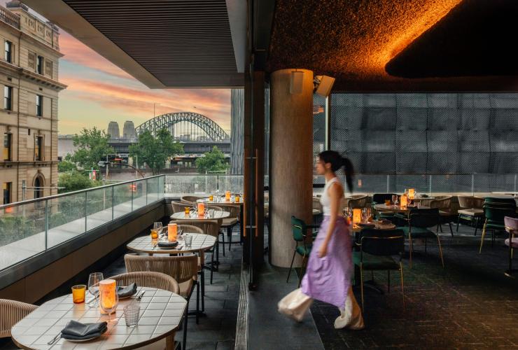 A person walking across the balcony of a restaurant with candle-lit tables overlooking a view of a large, steel arch bridge during a colourful pink sunset. Penelope's, Sydney, New South Wales © Penelope's/Chad Konik A person walking across the balcony of a restaurant with candle-lit tables overlooking a view of a large, steel arch bridge during a colourful pink sunset. Penelope's, Sydney, New South Wales © Penelope's/Chad Konik