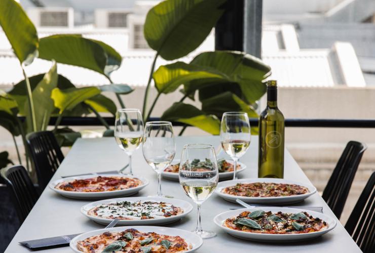 Une table près d'une fenêtre et d'une plante verte et feuillue avec un assortiment de pizzas et des verres de vin blanc au Gallery Bar, Adélaïde, Australie du Sud © Lewis Potter/Gallery Bar Une table près d'une fenêtre et d'une plante verte et feuillue avec un assortiment de pizzas et des verres de vin blanc au Gallery Bar, Adélaïde, Australie du Sud © Lewis Potter/Gallery Bar