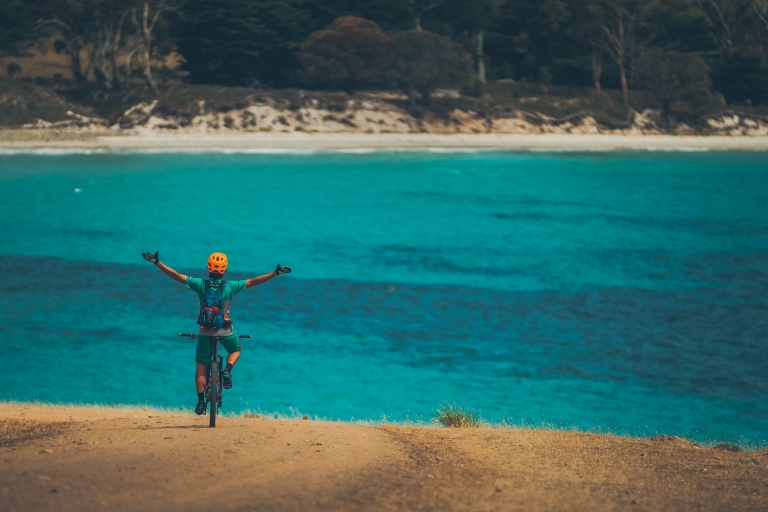 Une personne sur un VTT, bras levés en l'air, surplombant l'océan limpide dans le Maria Island National Park, Tasmanie © Matt Staggs