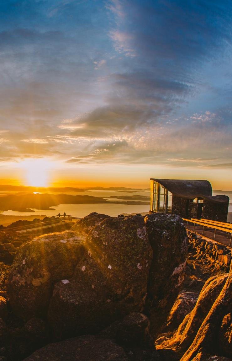 De gros rochers au premier plan avec deux personnes debout au loin sur un belvédère, admirant la vue sur Hobart, tandis que les rayons du soleil baignent le paysage du kunanyi/Mont Wellington, Hobart, Tasmanie © Tourism Australia
