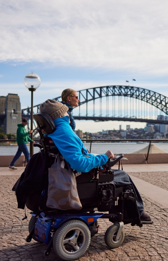 Orang yang menggunakan alat bantu mobilitas bersama orang lain yang berjalan di sampingnya mendekati Sydney Opera House dengan Sydney Harbour Bridge di latar belakangnya, di Sydney, New South Wales © Destination NSW