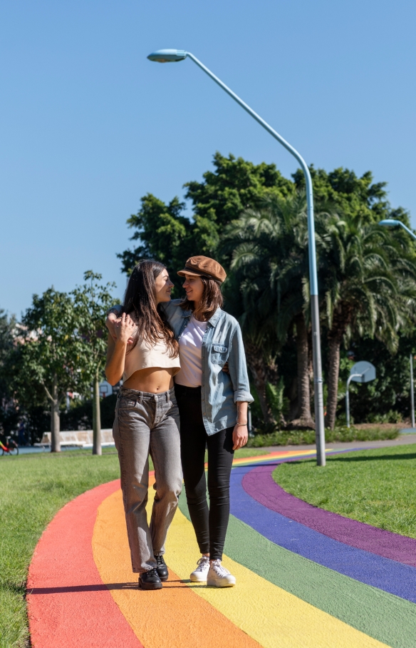 Pasangan sedang berjalan berangkulan menelusuri jalur pelangi di antara rumput hijau dan pepohonan di Prince Alfred Park, Surry Hills, New South Wales © Destination NSW