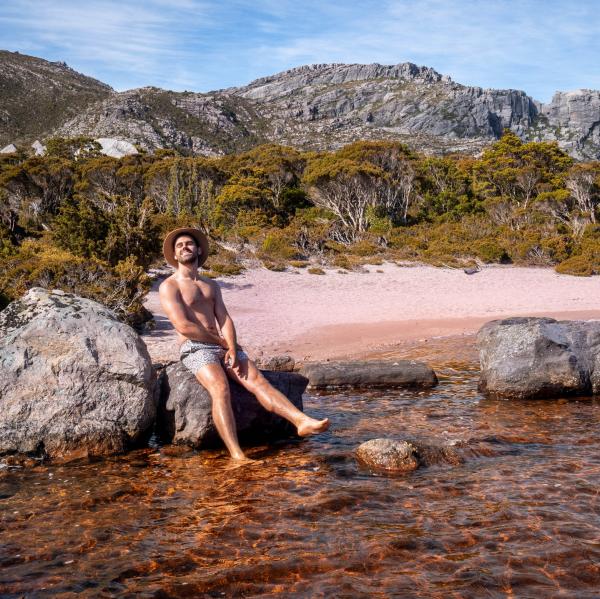 Miles Gray berpose di atas batu di sebuah danau di West-Coast Tasmania © Miles Gray, @milesuncharted