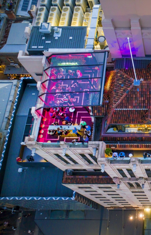 Pemandangan udara di atas Hennessy Rooftop Bar yang diterangi lampu merah muda dan dipenuhi orang-orang menikmati minuman di rooftop Mayfair Hotel, Adelaide, South Australia © Mayfair Hotel