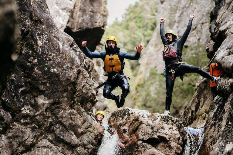 Dua orang memakai helm dan rompi pelampung di atas air terjun, salah satunya melompat dari bebatuan sementara yang lain bersorak di Cradle Mountain Canyons, Cradle Mountain, Tasmania © Tourism Australia