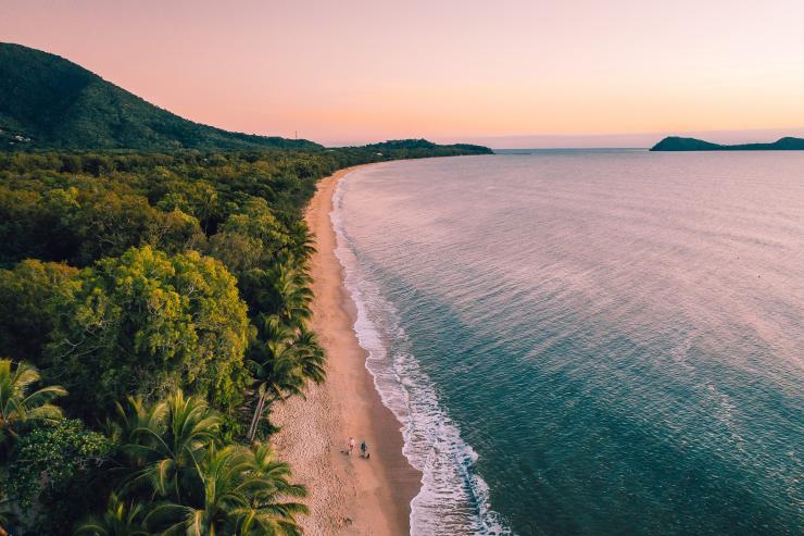 Pemandangan dari udara dua orang berjalan kaki di sepanjang hamparan pasir Clifton Beach di antara hutan hujan dan samudra saat matahari terbenam di Cairns, Queensland © Tourism and Events Queensland