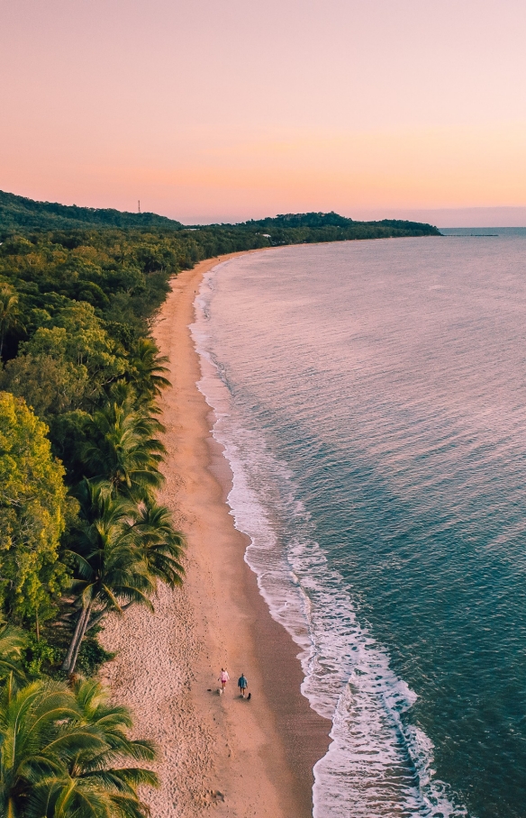 Pemandangan dari udara dua orang berjalan kaki di sepanjang hamparan pasir Clifton Beach di antara hutan hujan dan samudra saat matahari terbenam di Cairns, Queensland © Tourism and Events Queensland