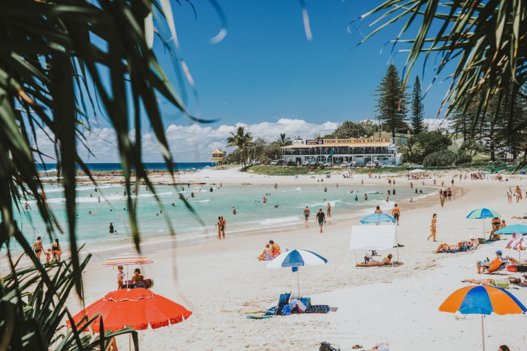 Orang-orang berbaring di pasir putih di bawah payung warna-warni sementara yang lainnya berenang di ombak yang tenang dan air biru jernih di Greenmount Beach di Coolangatta, Queensland © Tourism and Events Queensland