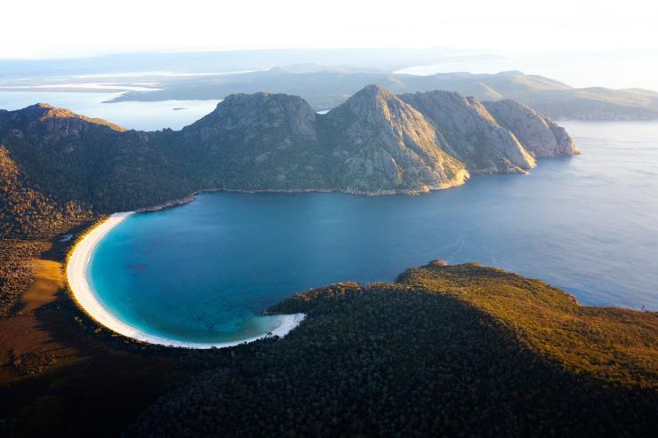 Pemandangan udara Wineglass Bay dan pegunungan di sekitarnya yang ditutupi pepohonan di Freycinet National Park, Tasmania © Lauren Bath