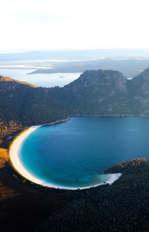 Pemandangan udara Wineglass Bay dan pegunungan di sekitarnya yang ditutupi pepohonan di Freycinet National Park, Tasmania © Lauren Bath