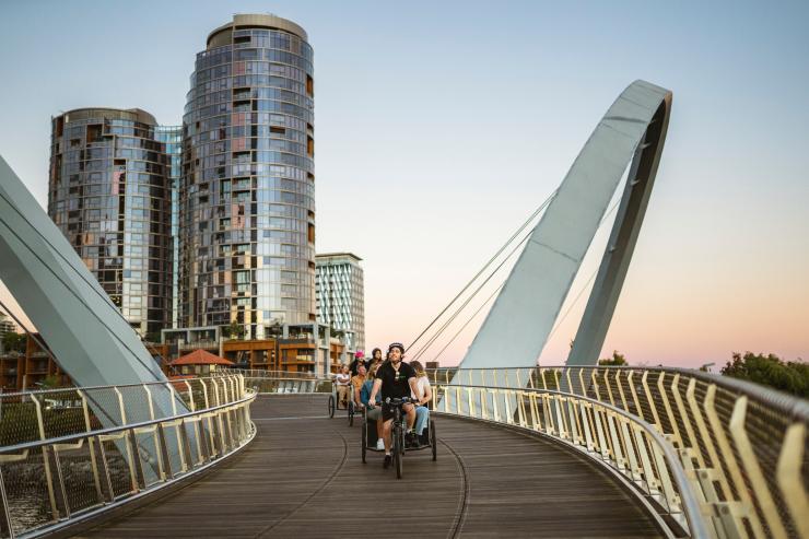 Sekelompok turis menikmati naik kereta roda dua melintasi jembatan di malam hari bersama Peddle Perth, Perth, Western Australia © Tourism Australia