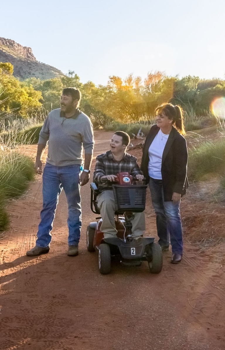 Uomo seduto sulla carrozzina di fianco ad un altro uomo e ad una donna ammirano la vegetazione lussureggiante e la sabbia rossa di Alice Springs, nell'Alice Spring Desert Park, Northern Territory.