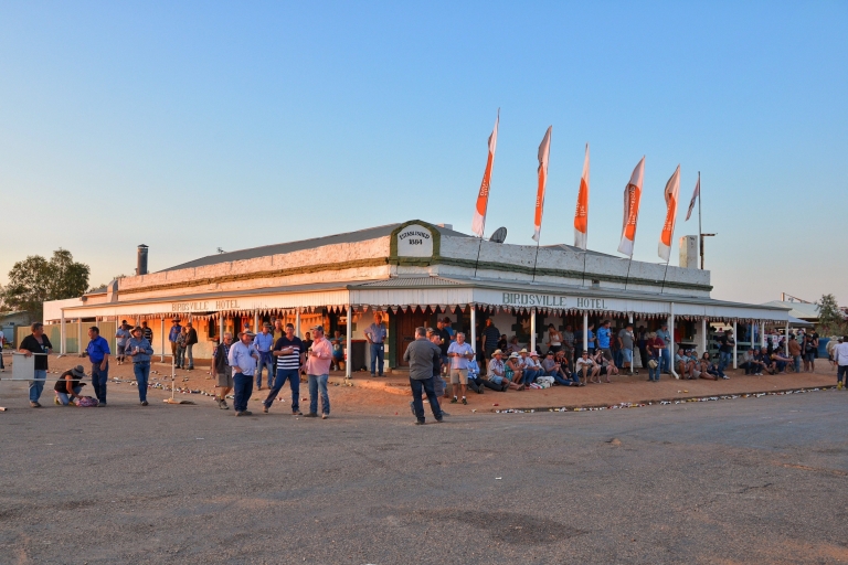 Storico pub con una tettoia decorata di bandiere e una folla di persone all'esterno, in piedi e sedute, che bevono e parlano al Birdsville Hotel, Birdsville, Queensland © Tourism and Events Queensland Storico pub con una tettoia decorata di bandiere e una folla di persone all'esterno, in piedi e sedute, che bevono e parlano al Birdsville Hotel, Birdsville, Queensland © Tourism and Events Queensland