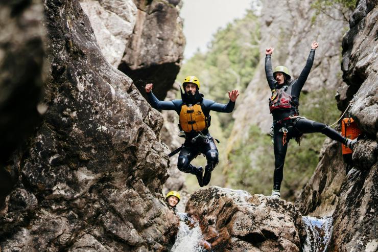 Due persone con caschetto e giubbotto salvagente in cima a una piccola cascata mentre uno salta dalla roccia e l'altro dietro gioisce a Cradle Mountain Canyons, Cradle Mountain, Tasmania © Tourism Australia