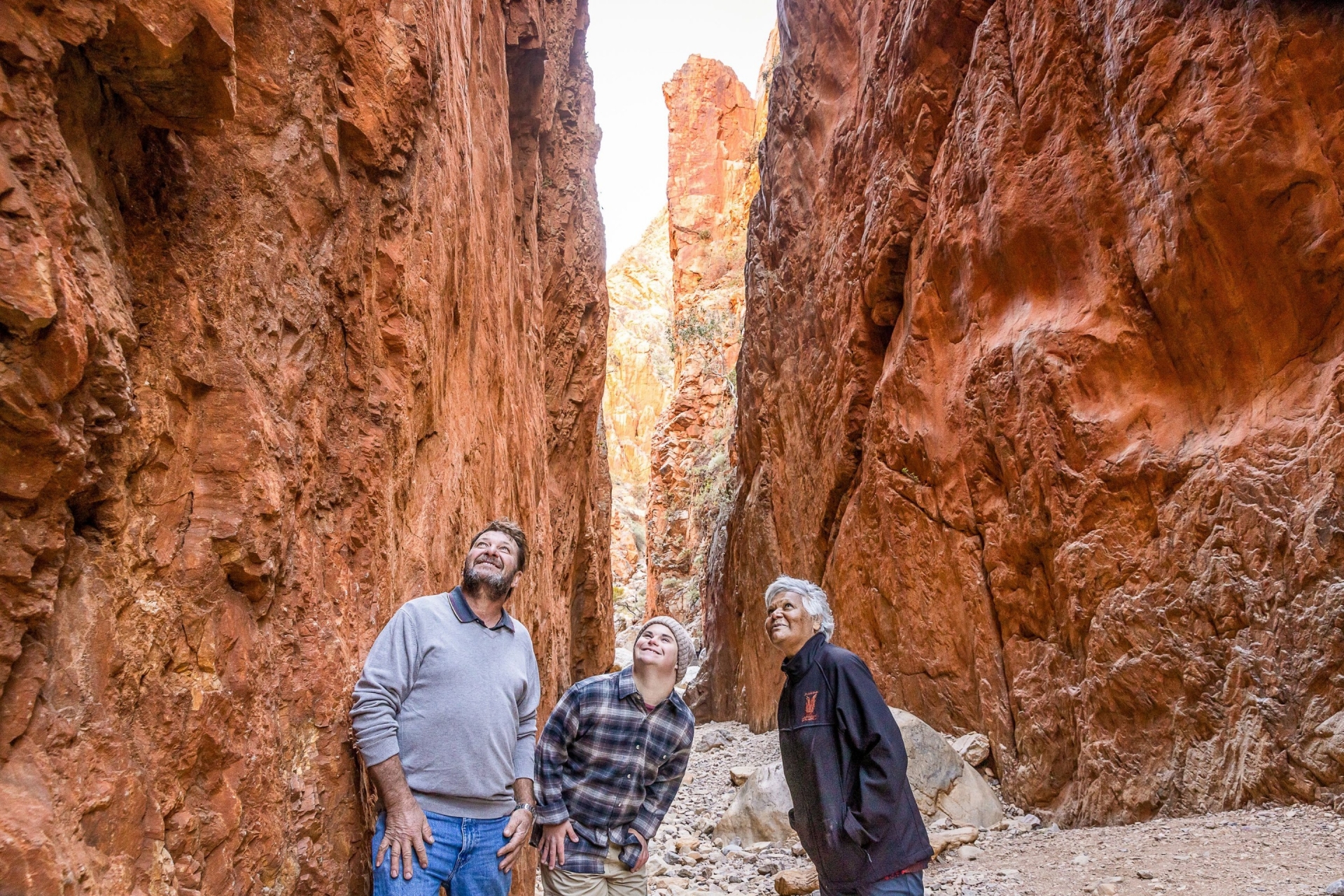 北領地西麥克唐奈爾山脈（West MacDonnell Ranges）斯坦德利裂縫（Standley Chasm）內一名神經多樣男子與另一名男子和一名導遊陪同©北領地旅遊局/Helen Orr