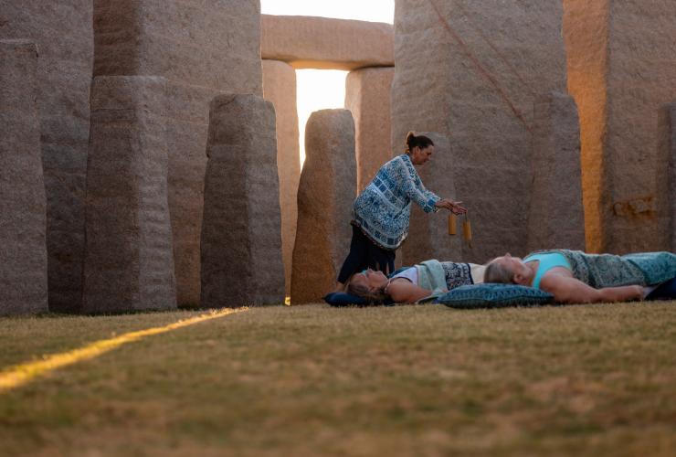 Gäste liegen auf Yogamatten und Kissen auf dem Gras inmitten von Steinstrukturen, während eine Lehrerin bei einem Retreat mit Untamed Escapes Instrumente über ihnen schwingt, Esperance, Westaustralien © Tourism Australia
