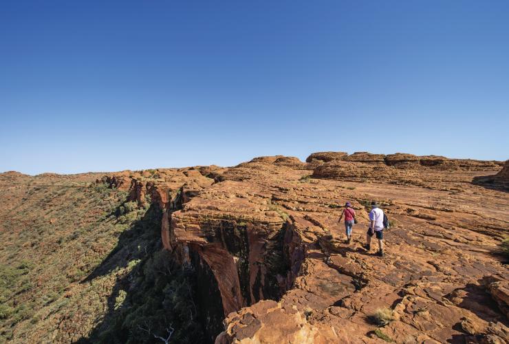 Zwei Menschen gehen am Rim Walk nah am Rand einer Klippe entlang, unter ihnen liegt Buschland, Kings Canyon, Northern Territory © Tourism Northern Territory/Shaana McNaught 