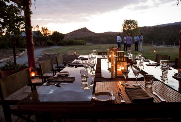 An elegant dining table set for a meal with candles illuminating the reflective table in the foreground, as a group of people stand on the grass in the distance drinking wine at Arkaba Conservancy, Flinders Ranges, South Australia © Arkaba Conservancy/Randy Larcombe