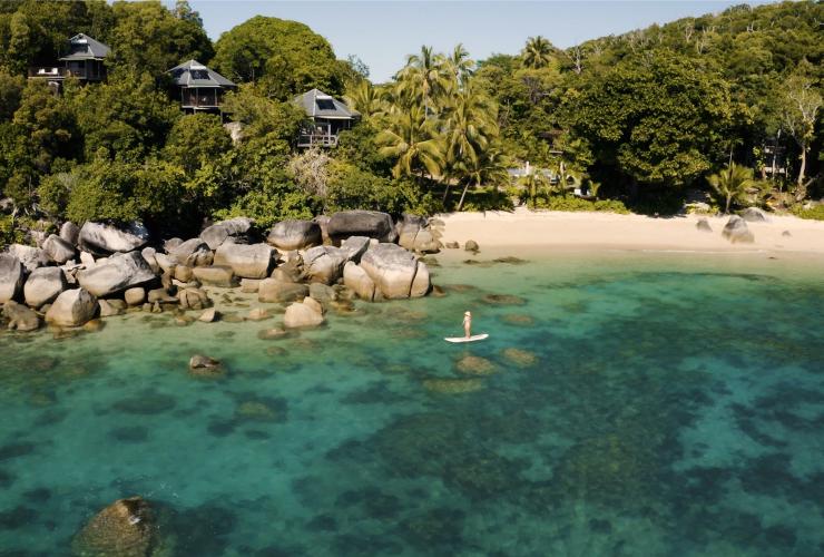 Aerial view over the coastline of Bedarra Island, with clear blue water, a sprinkling of large boulders, white sand and green palm trees on the Great Barrier Reef, Queensland © Tourism Australia