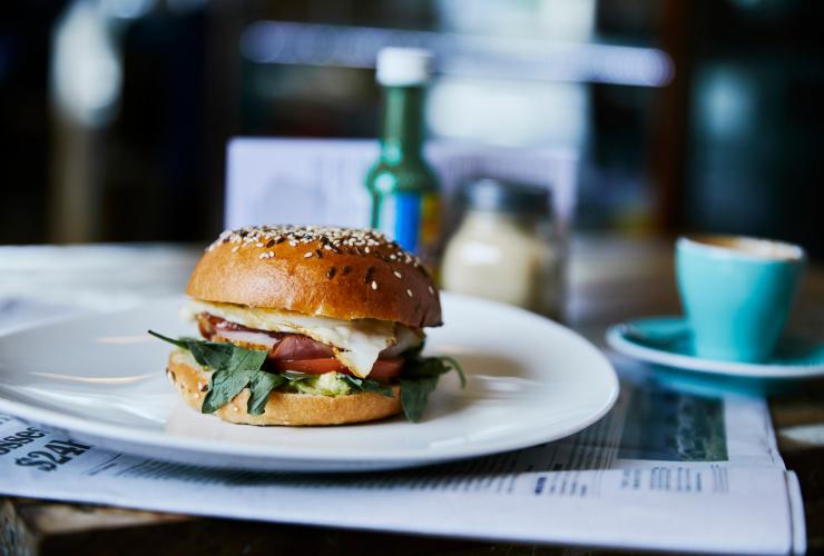 A bacon and egg roll in the foreground on a table with a mug and sauces behind it at Milking Station Cafe, Melbourne, Victoria © Visit Victoria