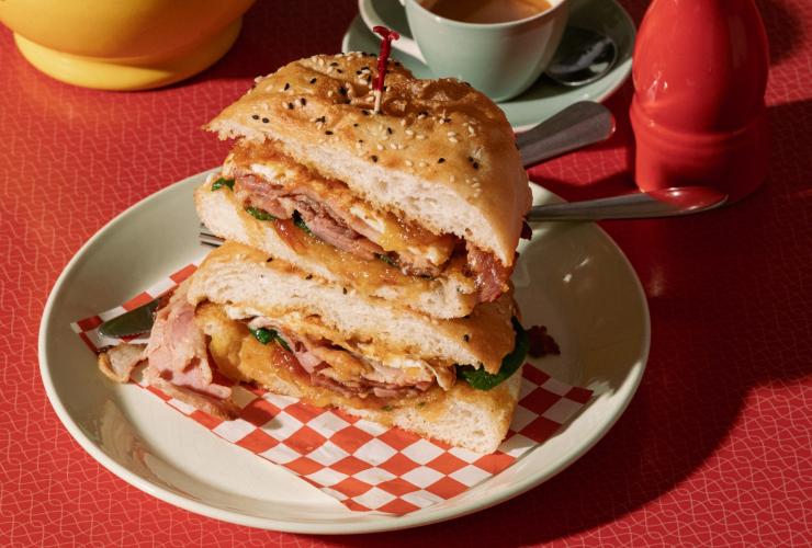 A bacon and egg roll cut into two halves stacked on each other beside a cup of coffee, a pepper shaker and a vase on a red table at Watervale General Store, Clare Valley, South Australia © South AUstralian Tourism Commission