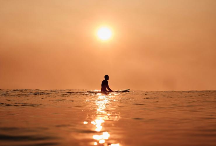 A surfer sits silouetted on a surfboard with an orange sunset behind them at Bondi Beach, Sydney, New South Wales © Tourism Australia