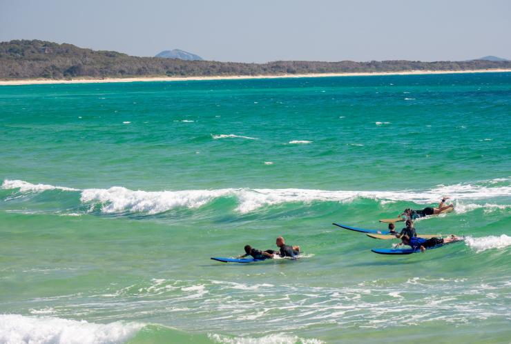 Several surfers lay on surfboards as a white-capped wave rolls in behind them at Noosa Main Beach, Noosa, Queensland © Tourism Australia