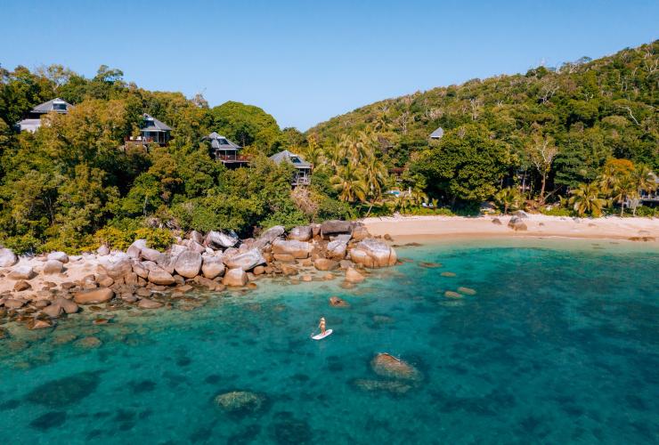 A person stand up paddle boarding in clear blue water with white sand shores leading to lush green rainforest behind them on Bedarra Island, Queensland © James Vodicka