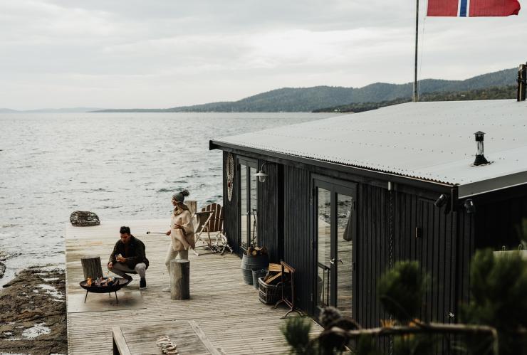 Two people gathered around an outdoor fire pit on a balcony beside a cottage elevated over the ocean at Satellite Island, Tasmania © Adam Gibson