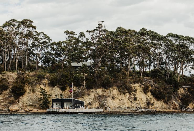 View across the calm blue ocean towards a seaside cottage beneath small rocky cliffs and native trees on Satellite Island, Tasmania © Adam Gibson