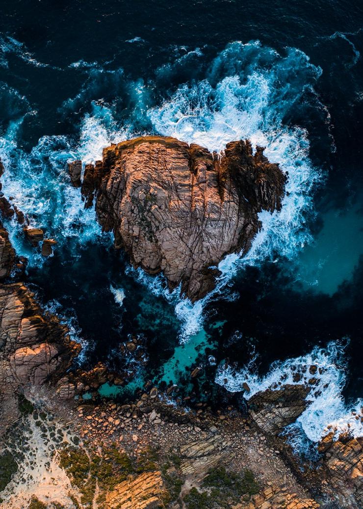 Aerial view over the round formation of Sugarloaf Rock surrounded by blue and white waves crashing against it in Margaret River, Western Australia © Tourism Australia