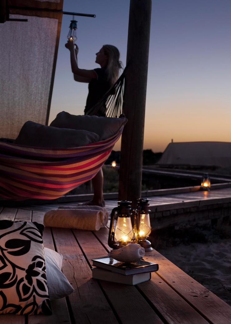 Lanterns, books and cushions sitting on a balcony in the foreground, with a woman standing beside a hammock and hanging a lantern during sunset at Sal Salis, Ningaloo Reef, Western Australia © Sal Salis Ningaloo Reef