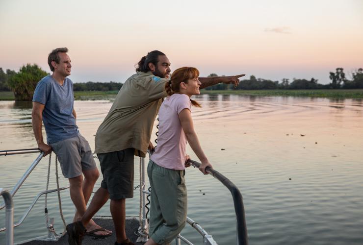 Two people standing with an Aboriginal guide as he points across a flat billabong reflecting the pastel colours of sunset during a Yellow Water Billabong cruise, Kakadu Tourism, Northern Territory © Tourism Australia