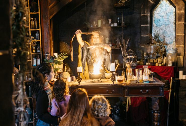 Parents et enfants regardant une actrice vêtue d'un costume d'enchanteresse et entourée d'accessoires préparant une potion explosive à Kryal Castle, Ballarat, Victoria © Tourism Australia/Visit Victoria