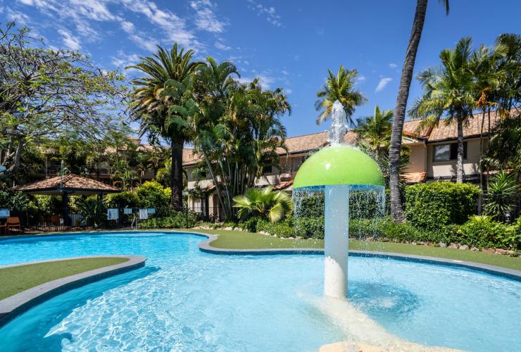 Air mancur di kolam renang yang dikelilingi pohon palem dengan bangunan di belakangnya di Turtle Beach Resort, Gold Coast, Queensland © Turtle Beach Resort