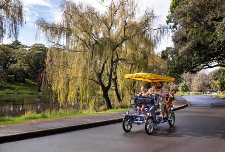 Famiglia su un risciò coperto da un tettuccio accanto a un laghetto tra la vegetazione del Centennial Park Cycles, Centennial Park, New South Wales © Destination NSW
