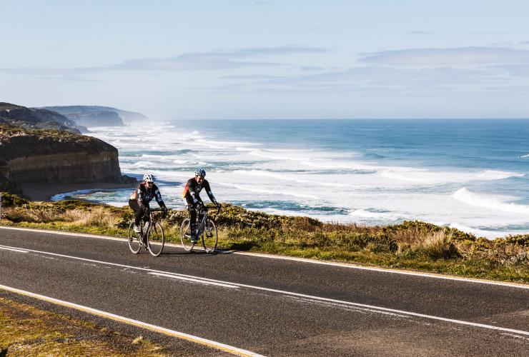 Due persone che percorrono in bicicletta una di fianco all'altra una strada accanto a ripide scogliere in arenaria a picco sull'oceano lungo la Great Ocean Road, Victoria © Belinda Van Zanen/Great Ocean Road Tourism
