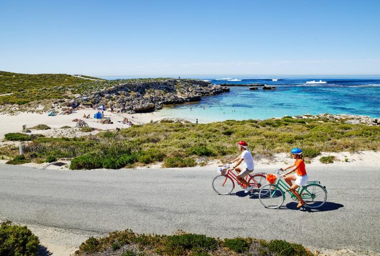 Due persone che pedalano su biciclette colorate lungo una strada accanto a una baia con sabbia bianca e acque cristalline a Little Salmon Bay, Rottnest Island, Western Australia © Tourism Western Australia