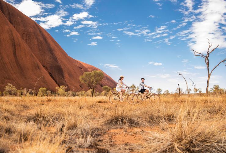 Due persone che pedalano in una pianura erbosa con le torreggianti pareti di roccia rossa di Uluru alle loro spalle nell'Uluru-Kata Tjuta National Park, Northern Territory © Tourism NT